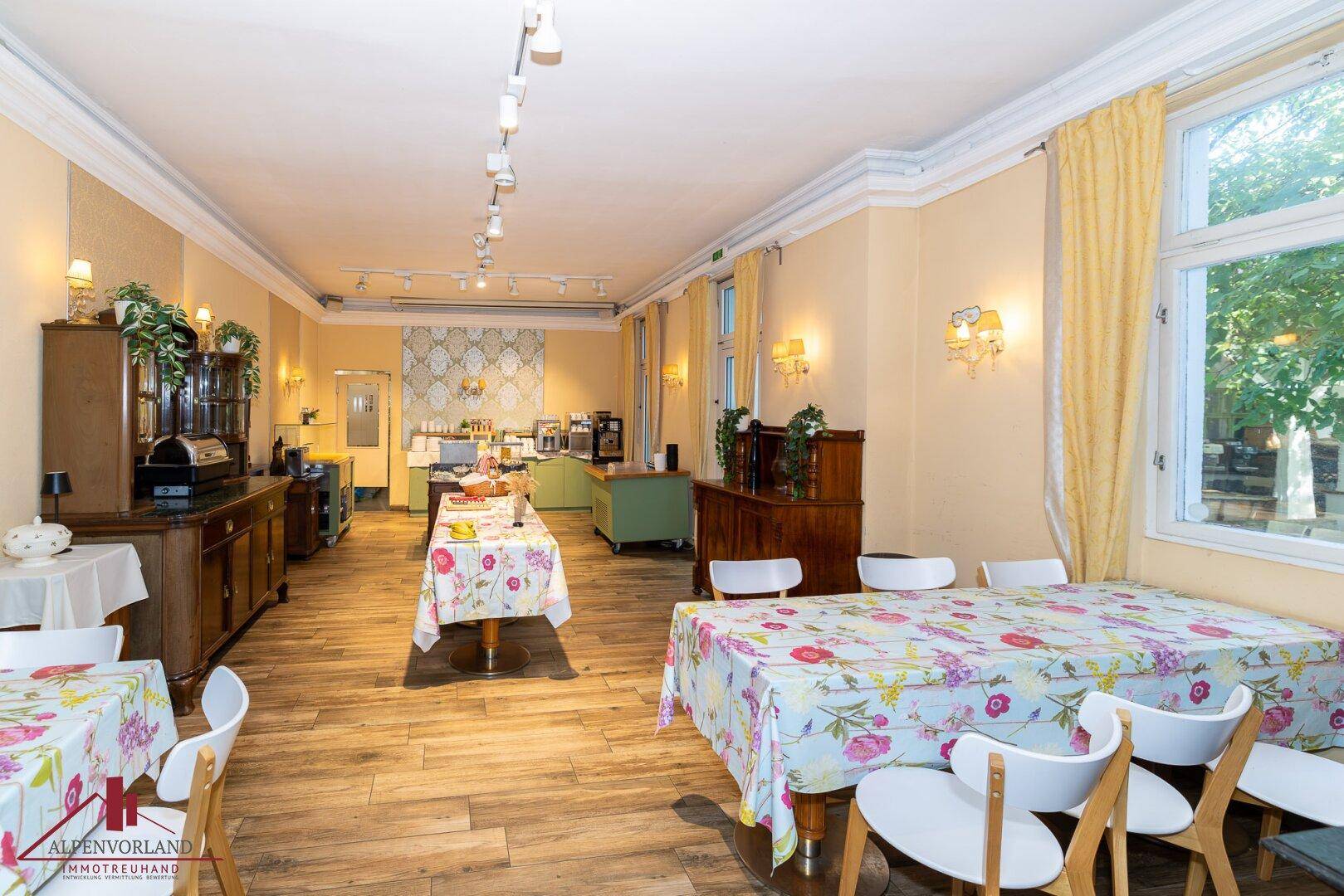 Heller Speisesaal mit Holzboden, großen Fenstern und floralen Tischdecken.