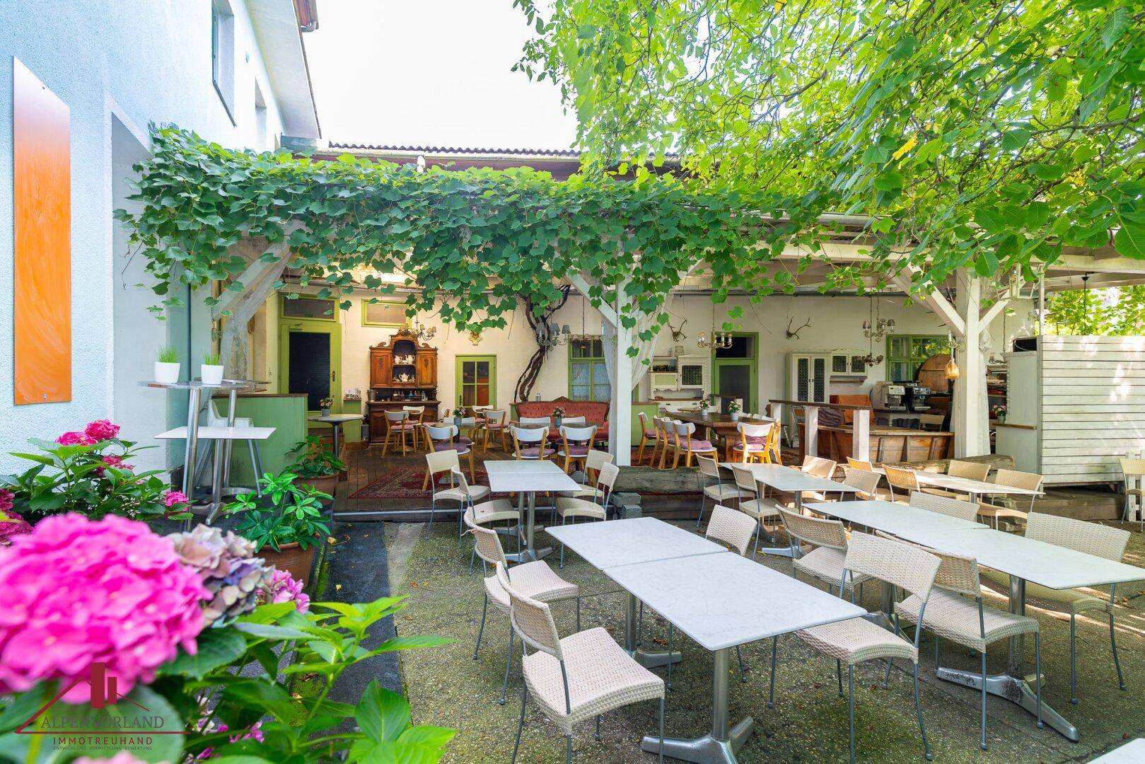 Grüne Außenterrasse mit vielen Sitzgelegenheiten unter einer bewachsenen Pergola.