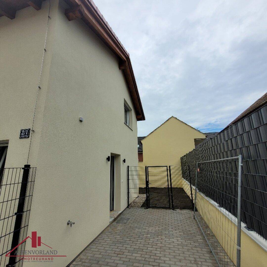 Seitenansicht des Hauses mit gepflastertem Zugangsweg und einem Zaun unter bewölktem Himmel.
