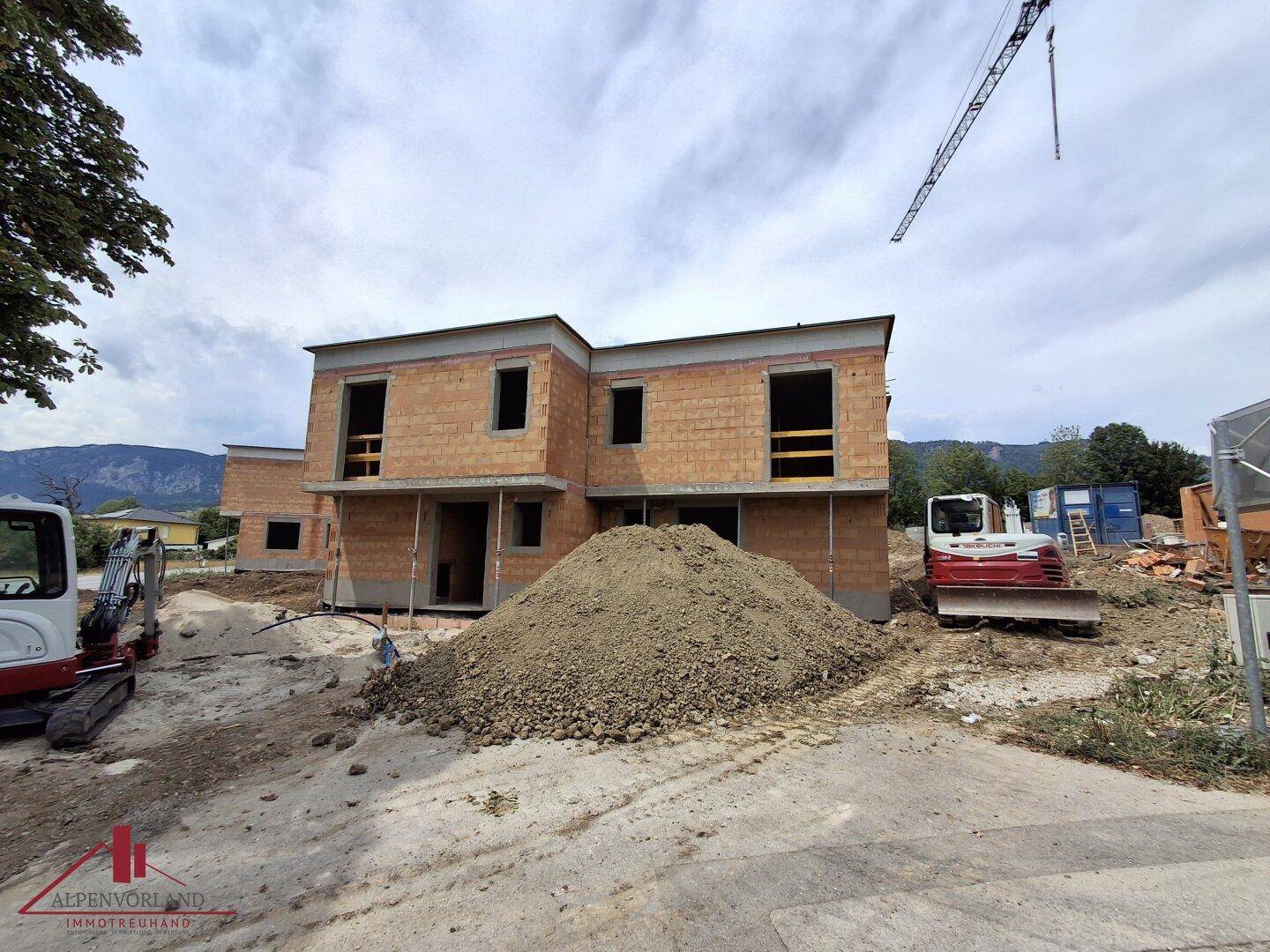 Baustelle mit zwei Rohbauten und einem Kran im Hintergrund, umgeben von Erdhügeln.