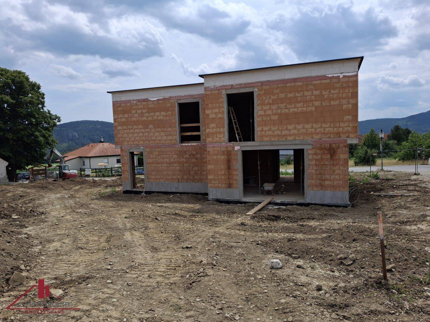 Baustelle mit zwei Rohbauten, umgeben von Erdhügeln und Bauschutt unter bewölktem Himmel.