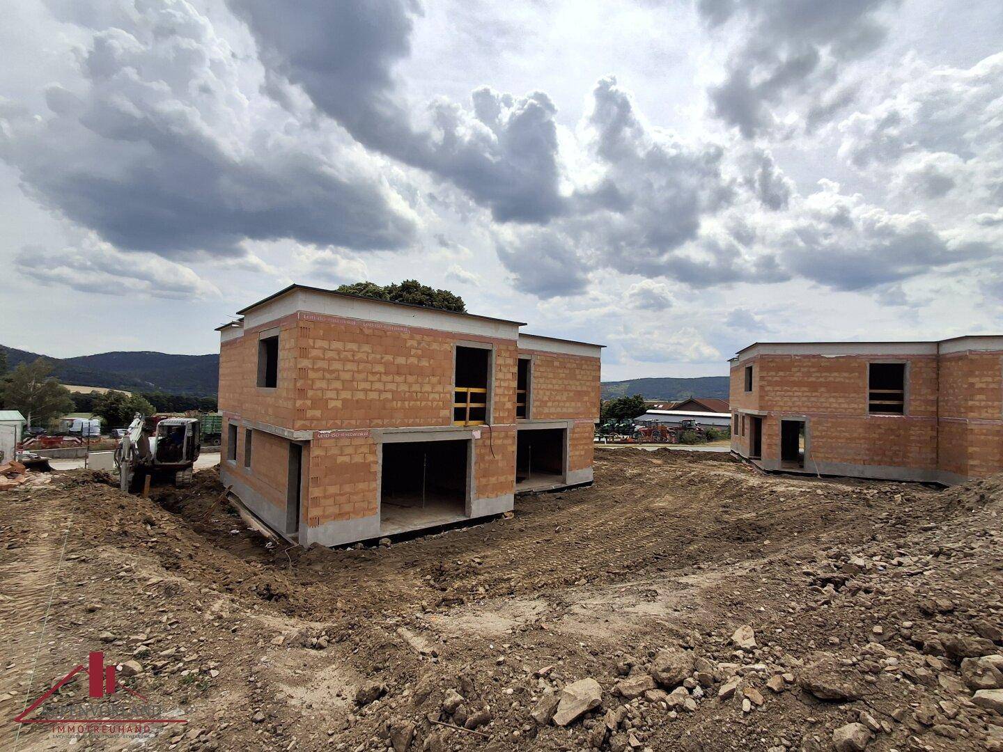 Weitläufige Ansicht der Baustelle mit zwei Rohbauten unter einem bewölkten Himmel und Hügeln im Hintergrund.