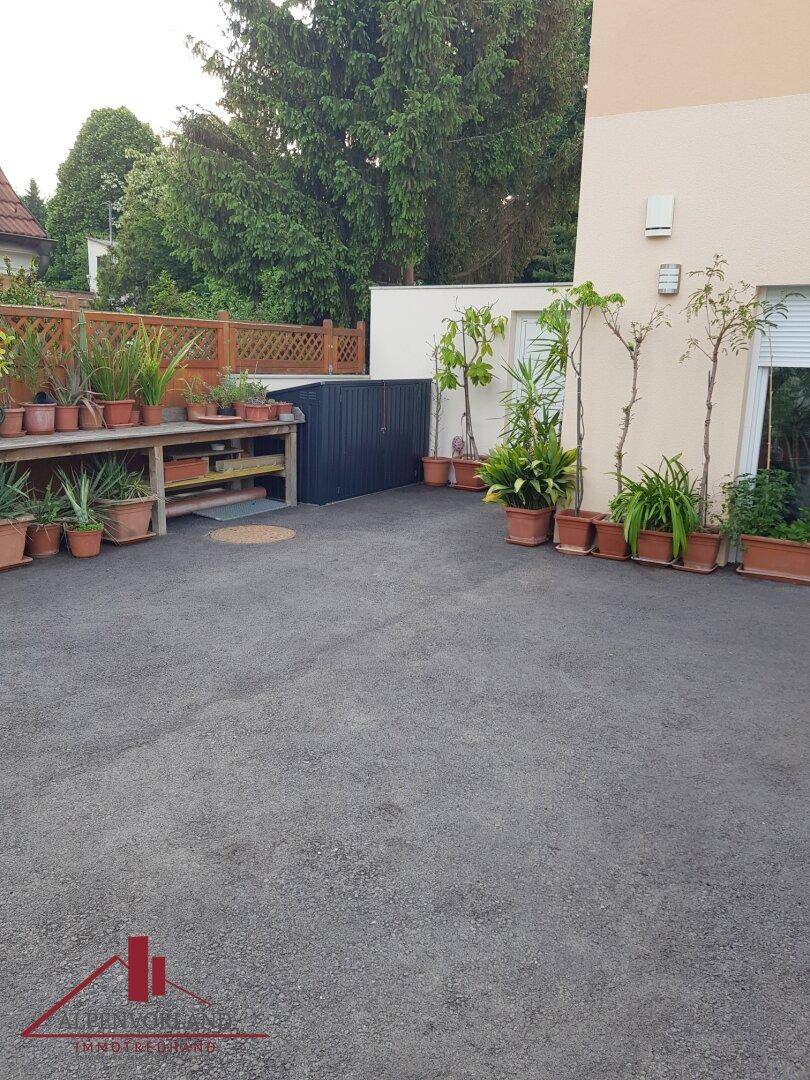Gepflasterter Außenbereich mit Topfpflanzen entlang der Hauswand und einem kleinen Abstellschuppen.