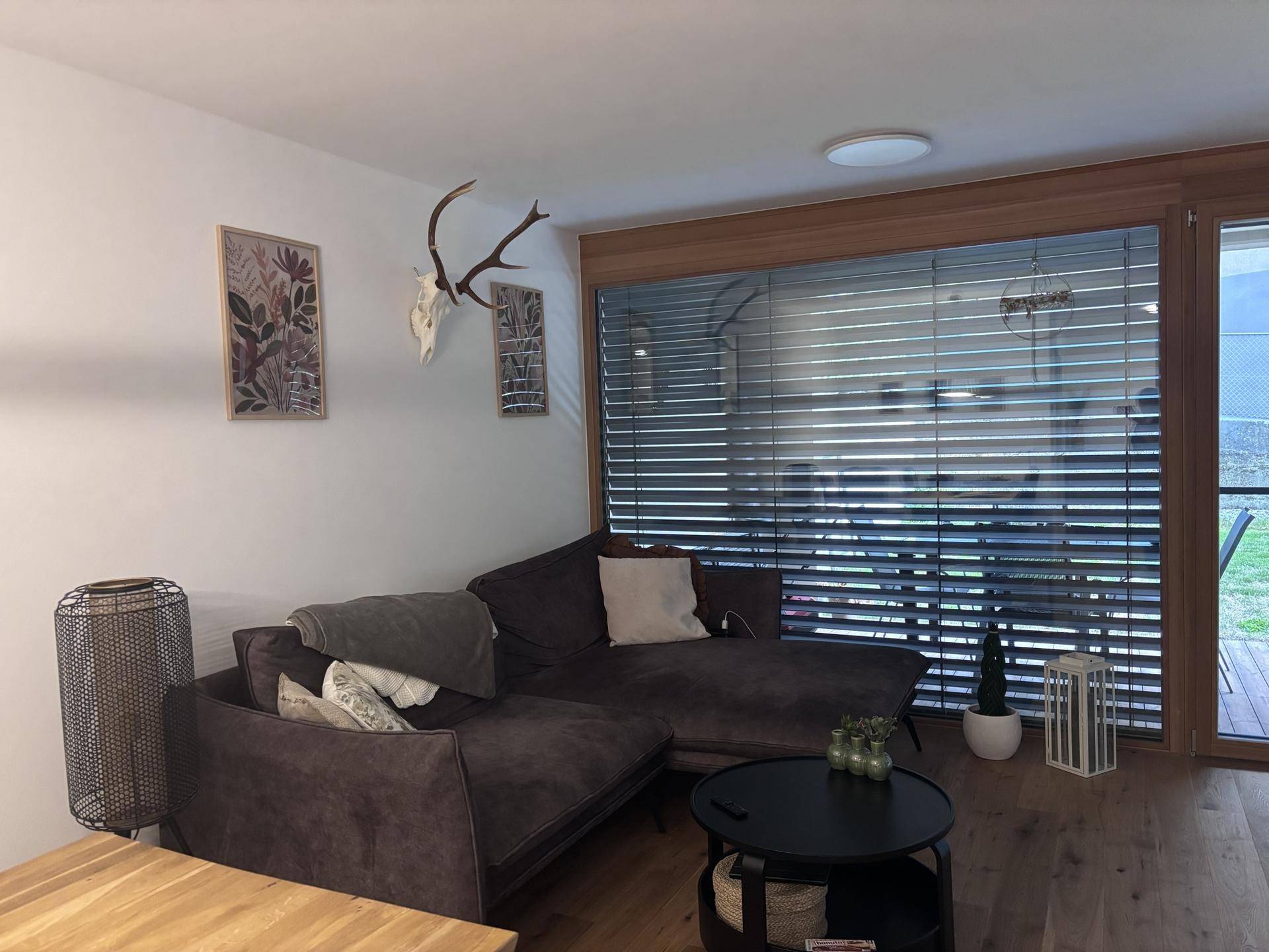 Gemütliches Wohnzimmer mit Ecksofa, Couchtisch und großen Fenstern mit Jalousien.