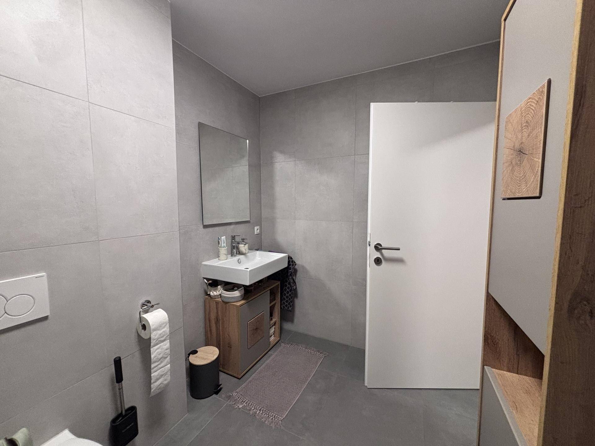 Badezimmer mit Einzelwaschbecken, Spiegel und WC, ausgestattet mit grauen Fliesen.