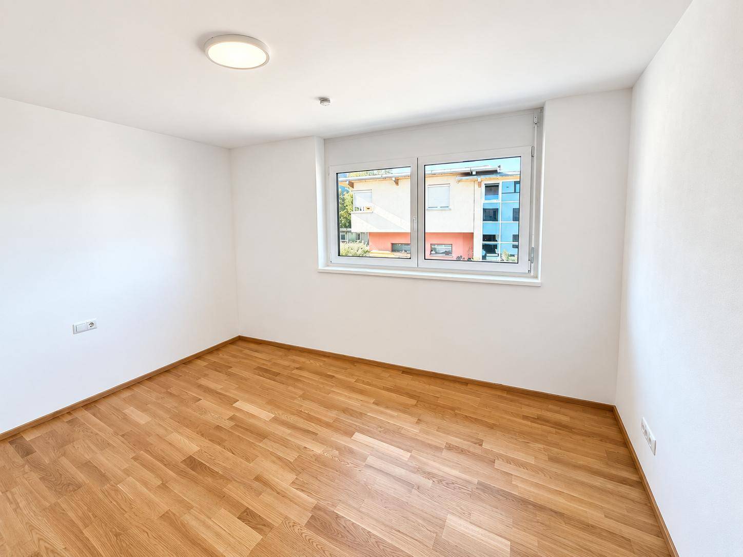 Helles Schlafzimmer mit Parkettboden und einem Fenster, das den Blick auf die Umgebung freigibt.