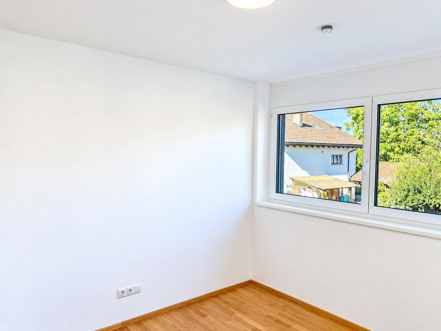 Helles Zimmer mit Holzboden und Fensterblick ins Grüne und auf Nachbarhäuser.