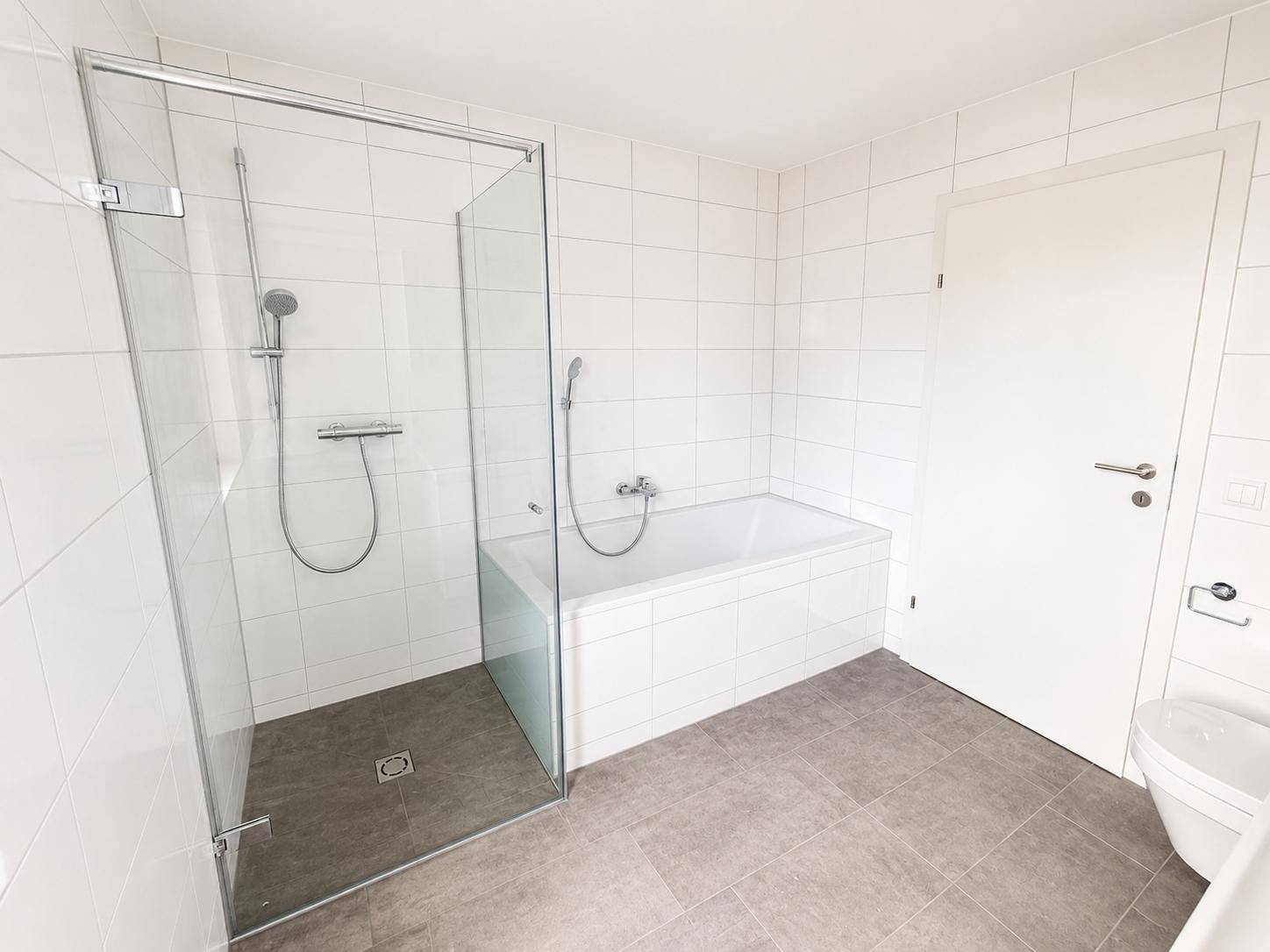 Modernes Badezimmer mit weißen Fliesen, begehbarer Dusche, Badewanne und grauen Bodenfliesen.