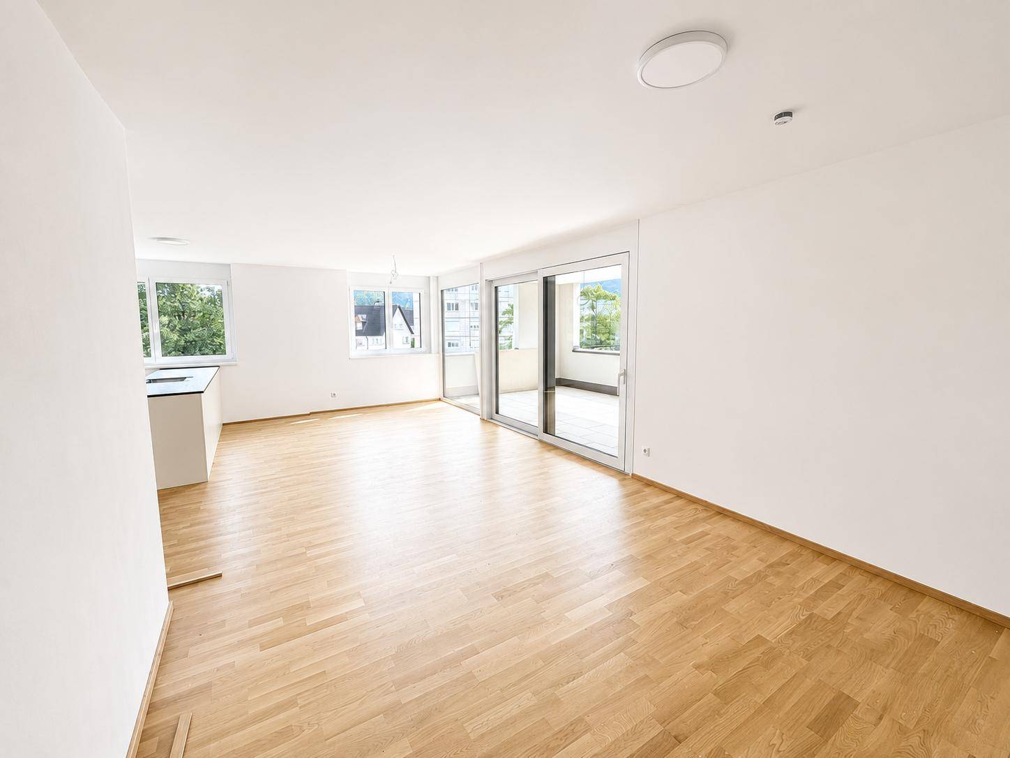 Heller, leerer Wohnbereich mit Parkettboden, großen Fenstern und Zugang zu einem Außenbereich.
