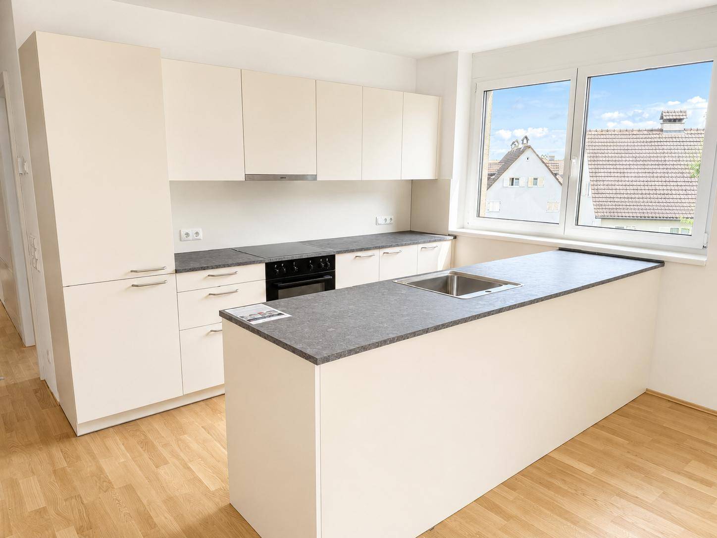 Moderne Einbauküche mit hellen Schränken, dunkler Arbeitsplatte und Fenster mit Blick ins Grüne.