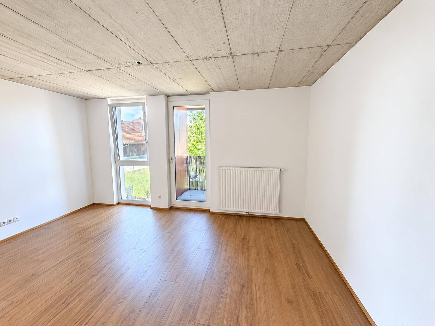 Geräumiges Wohnzimmer mit viel Tageslicht, Laminatboden und moderner Sichtbetondecke.