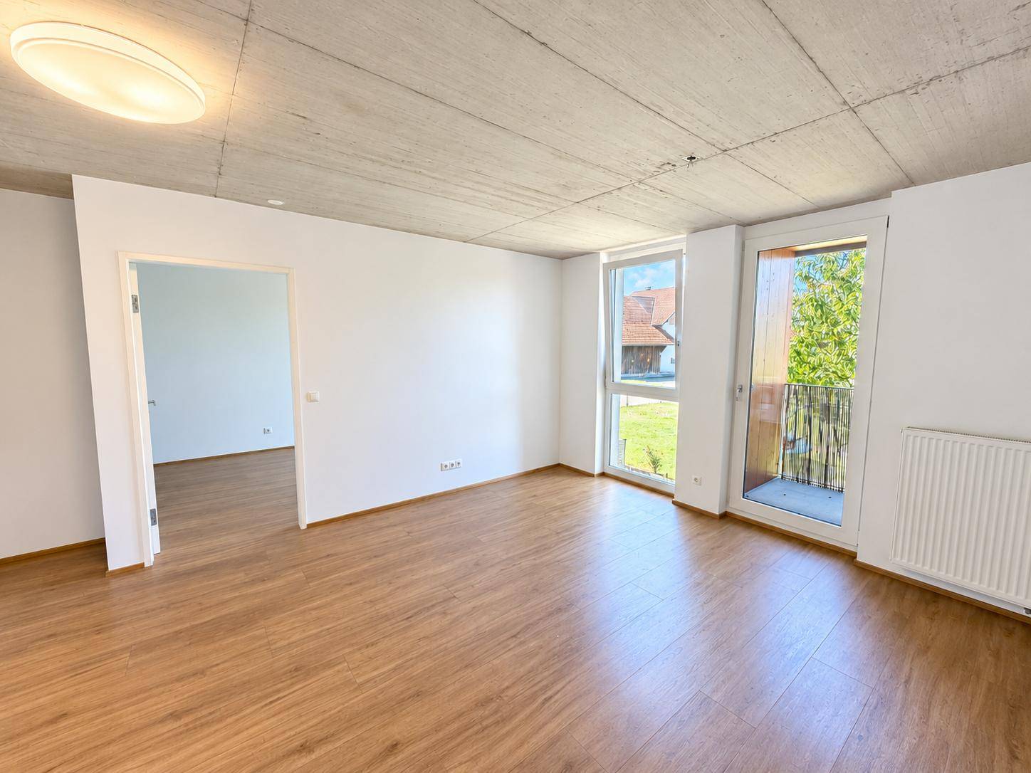 Helles Wohnzimmer mit Laminatboden, Sichtbetondecke und Zugang zu einem französischen Balkon.