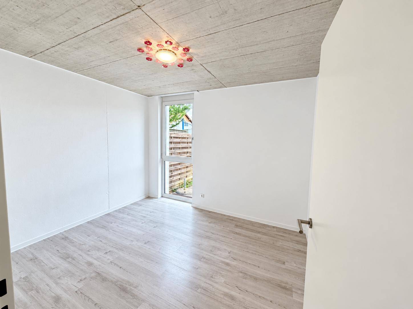 Helles Schlafzimmer mit Laminatboden, Sichtbetondecke und Fenster mit Blick ins Grüne.