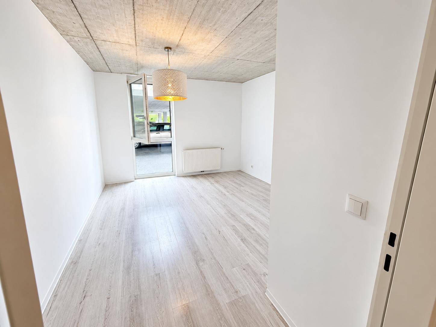 Helles Zimmer mit Laminatboden, Sichtbetondecke und Zugang zu einem kleinen Außenbereich.
