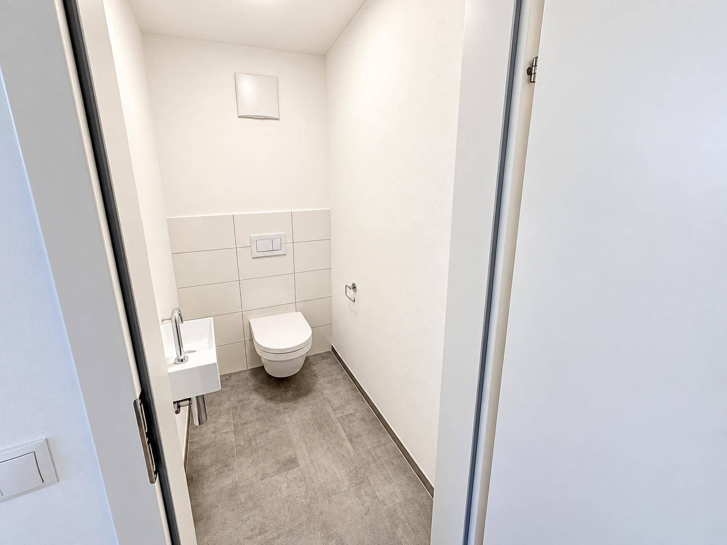 Kleines Gäste-WC mit Wand-WC, Handwaschbecken und weißen Fliesen im unteren Bereich.