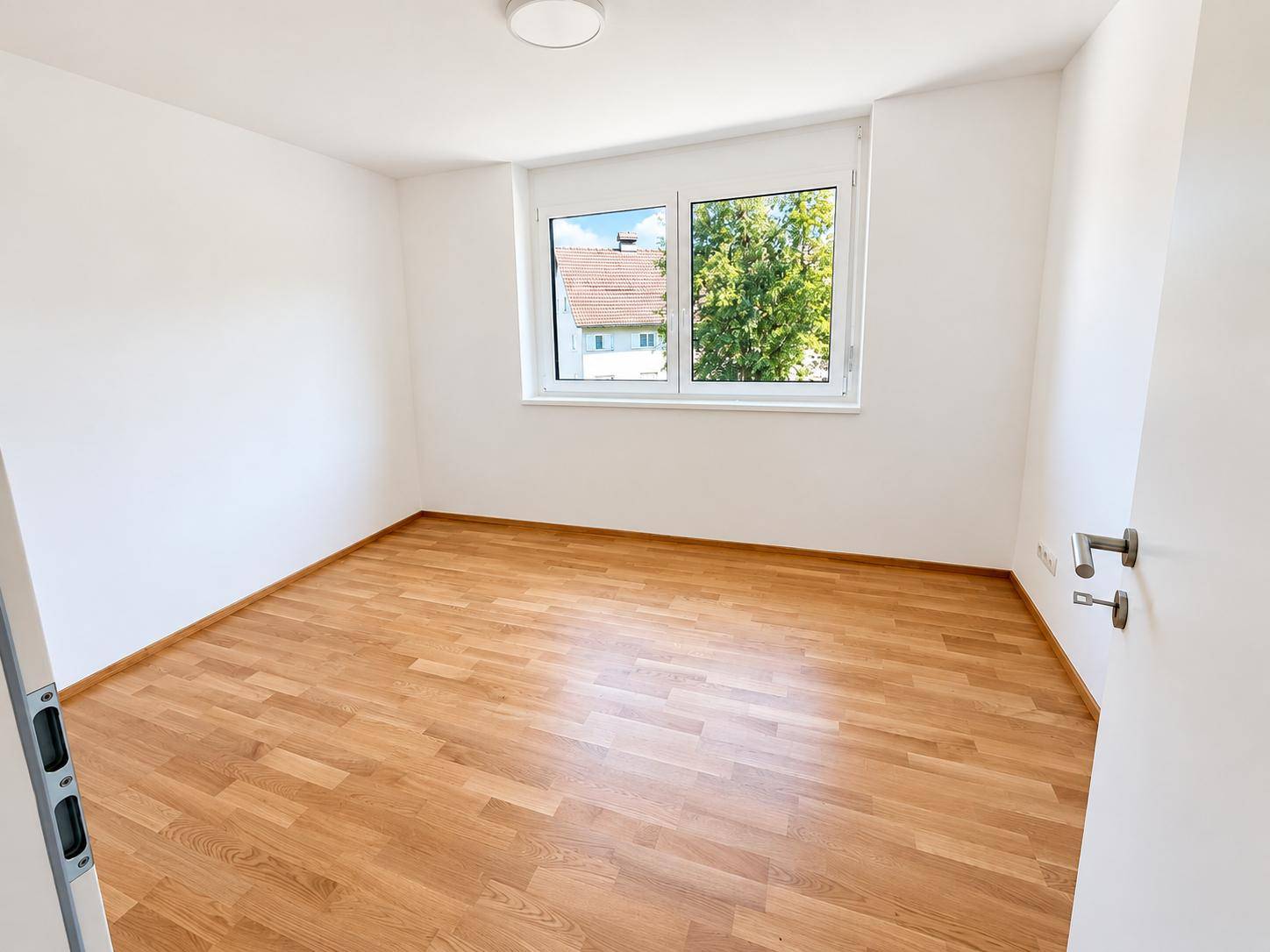 Helles Schlafzimmer mit Holzparkettboden und einem Fenster mit Blick ins Grüne.