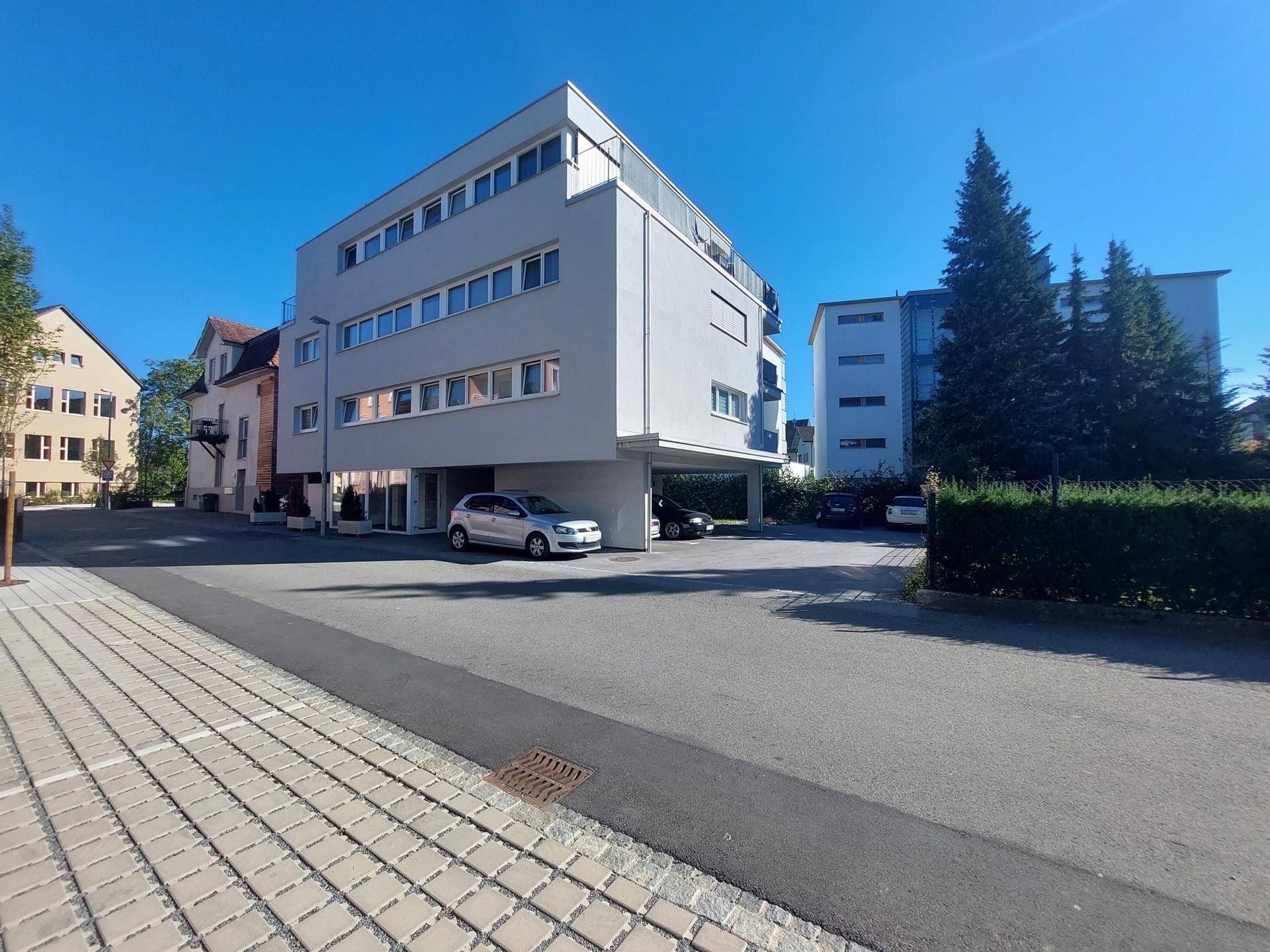 Ansicht eines modernen Wohngebäudes mit hellgrauer Fassade, Carport und umliegender Grünfläche an einem sonnigen Tag.