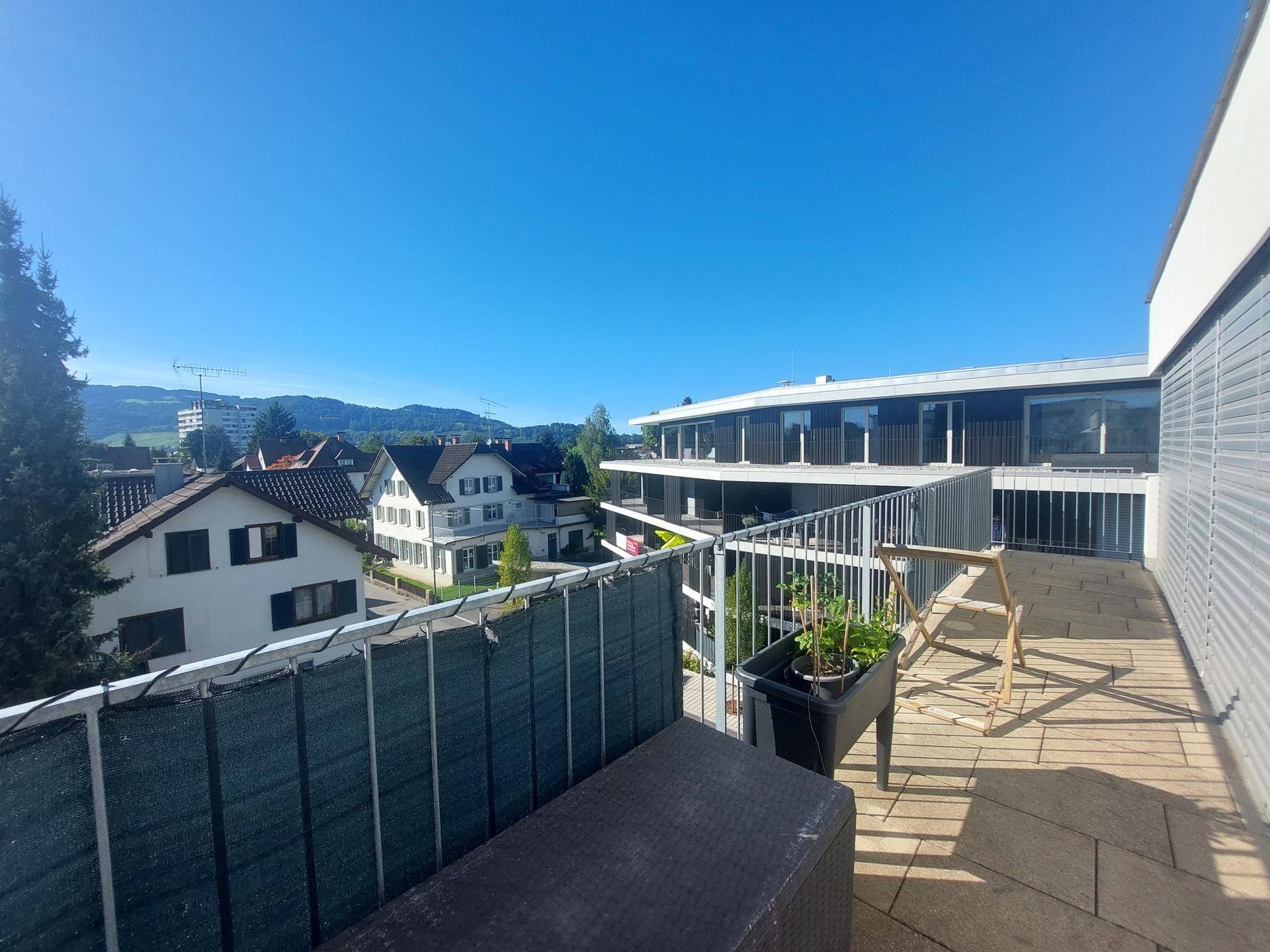 Breiter Balkon mit Blick über die Dächer der Nachbarschaft und die umliegende Landschaft unter klarem Himmel.