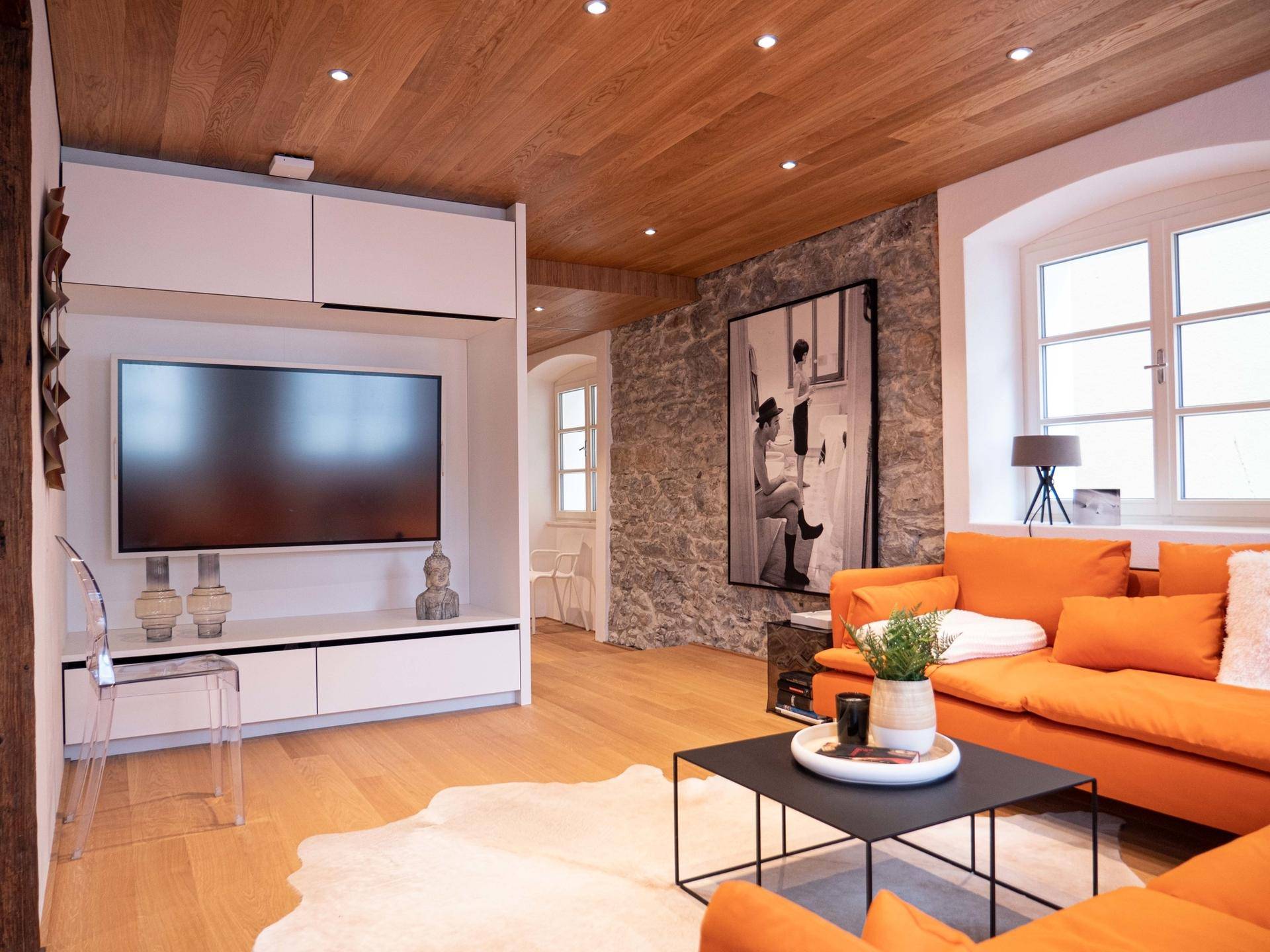 Gemütlicher Wohnbereich mit orangefarbenem Sofa, Holzbalkendecke und rustikaler Steinwand.