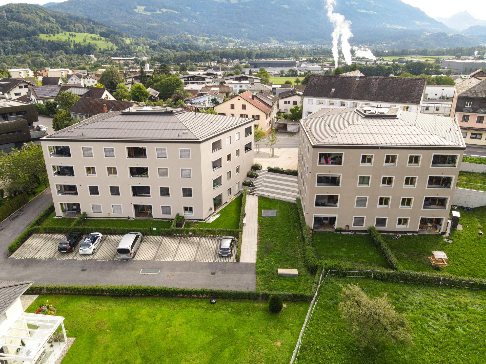 Luftaufnahme der beiden modernen Wohngebäude mit Parkplätzen und einem Weg dazwischen, vor einer Bergkulisse.