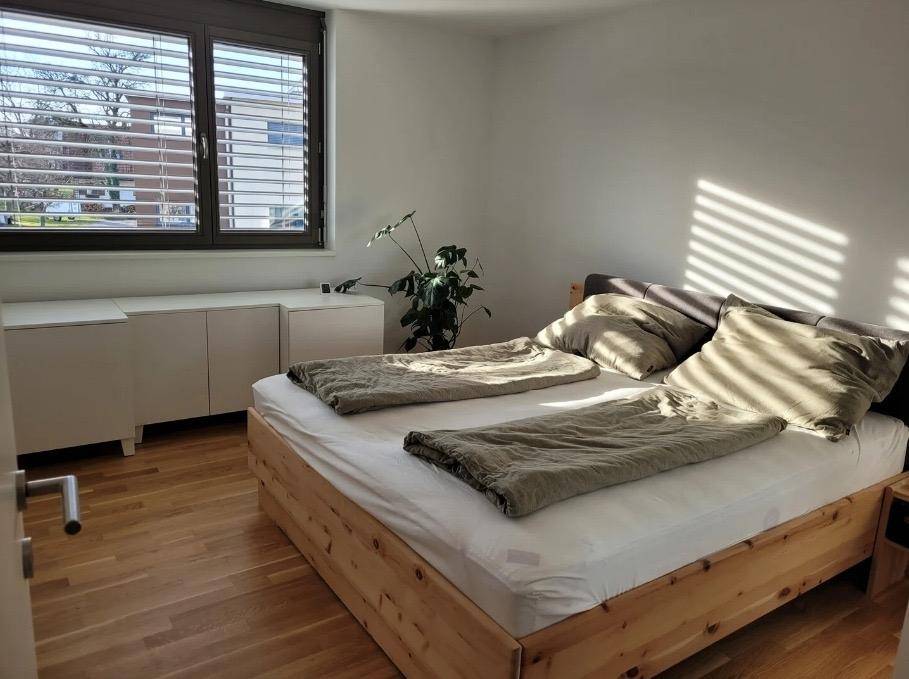 Sonniges Schlafzimmer mit Holzbett und breitem Fenster, das viel Tageslicht hereinlässt.