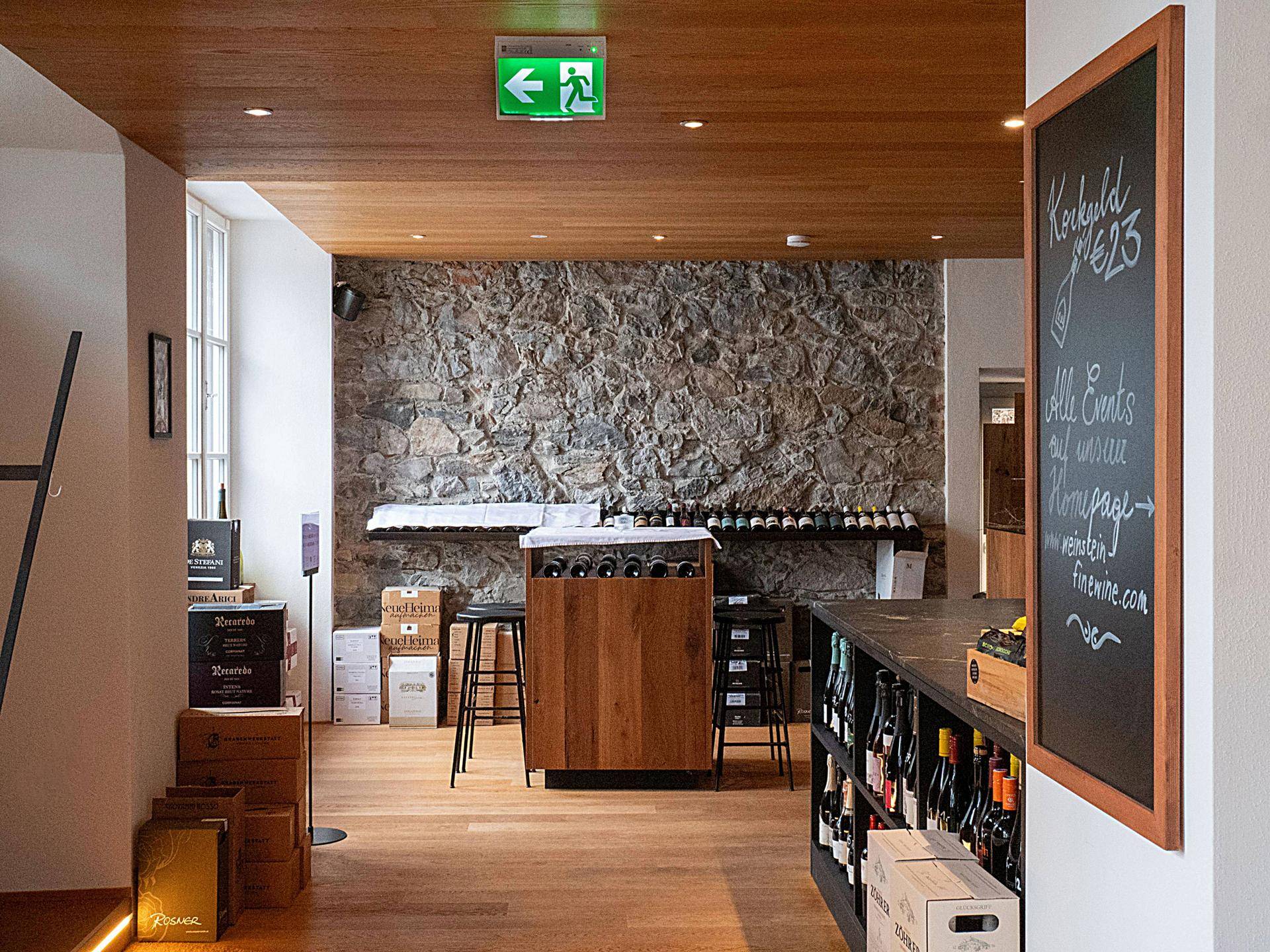 Heller Verkaufsraum mit Holzboden, rustikaler Steinwand und modernen Barhockern.