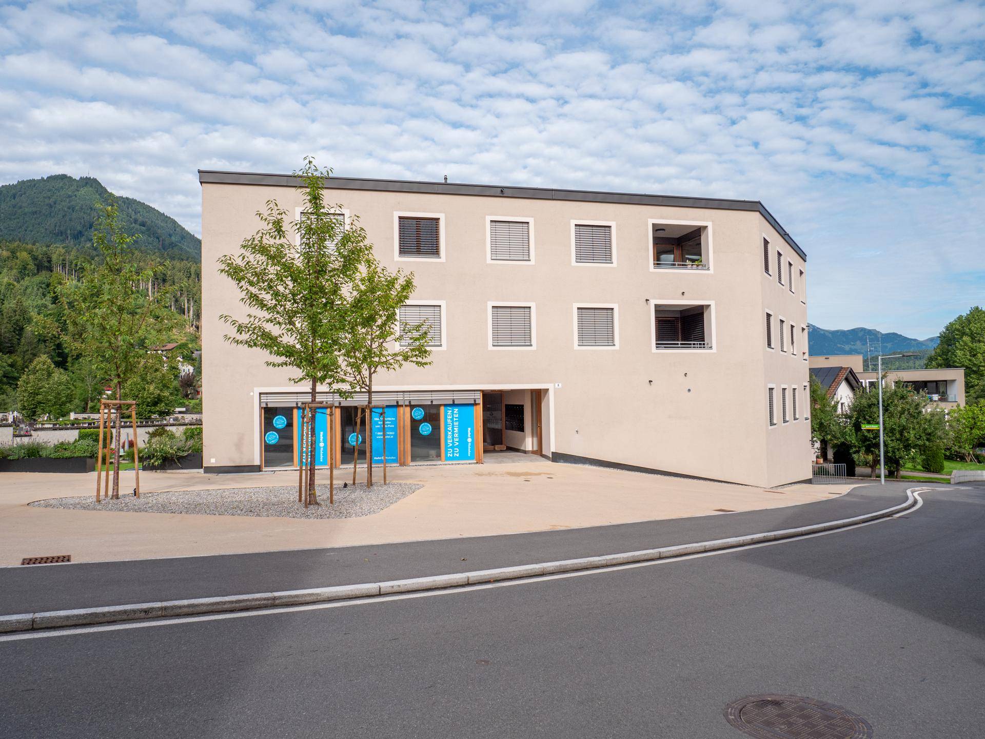 Vorderansicht eines modernen Gebäudes mit beiger Fassade, Fenstern mit Rollläden und Geschäftsräumen im Erdgeschoss, mit Bergblick.