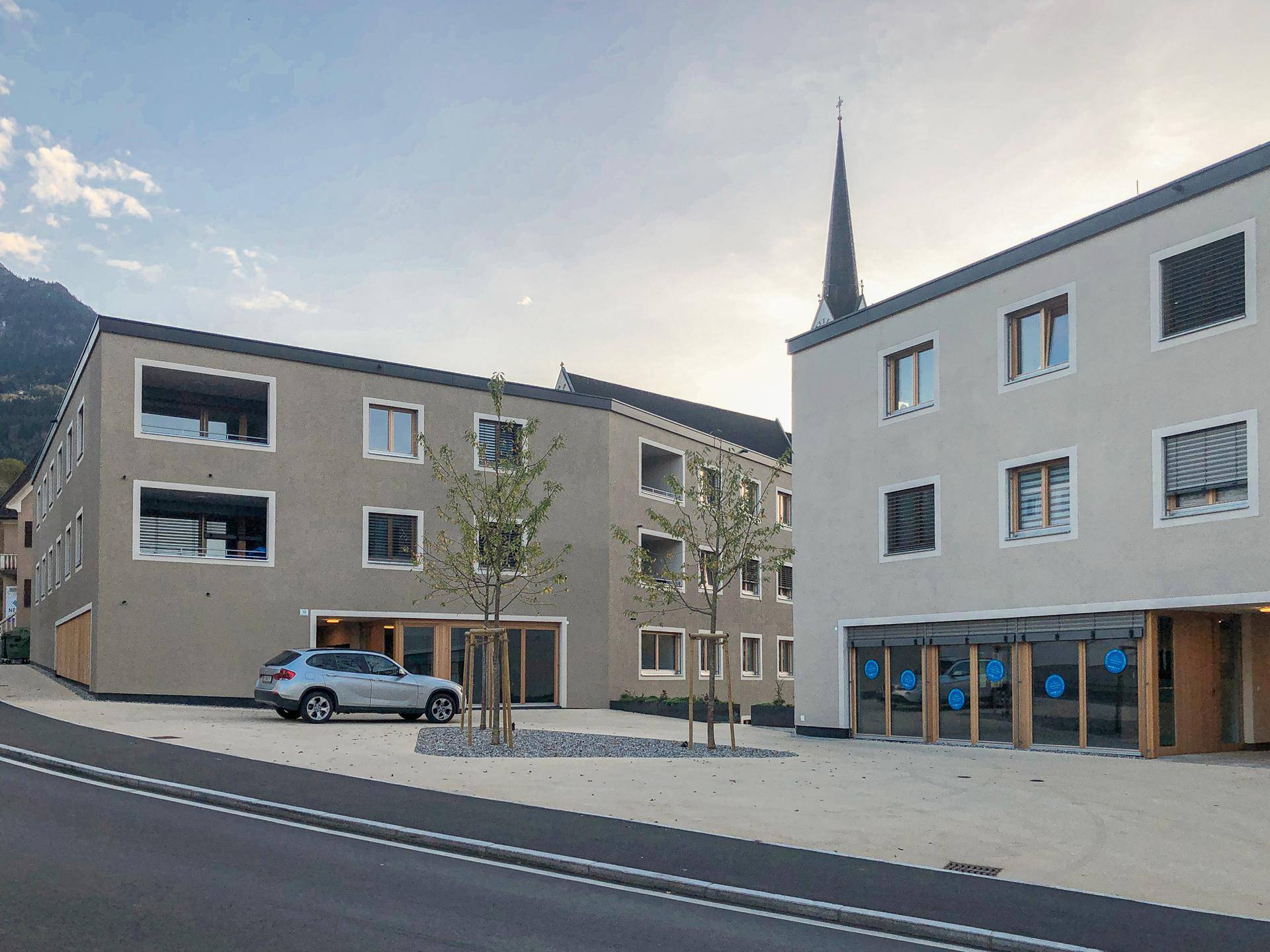 Straßenansicht der beiden modernen Gebäude in der Dämmerung, mit einem geparkten Auto und einem Kirchturm im Hintergrund.