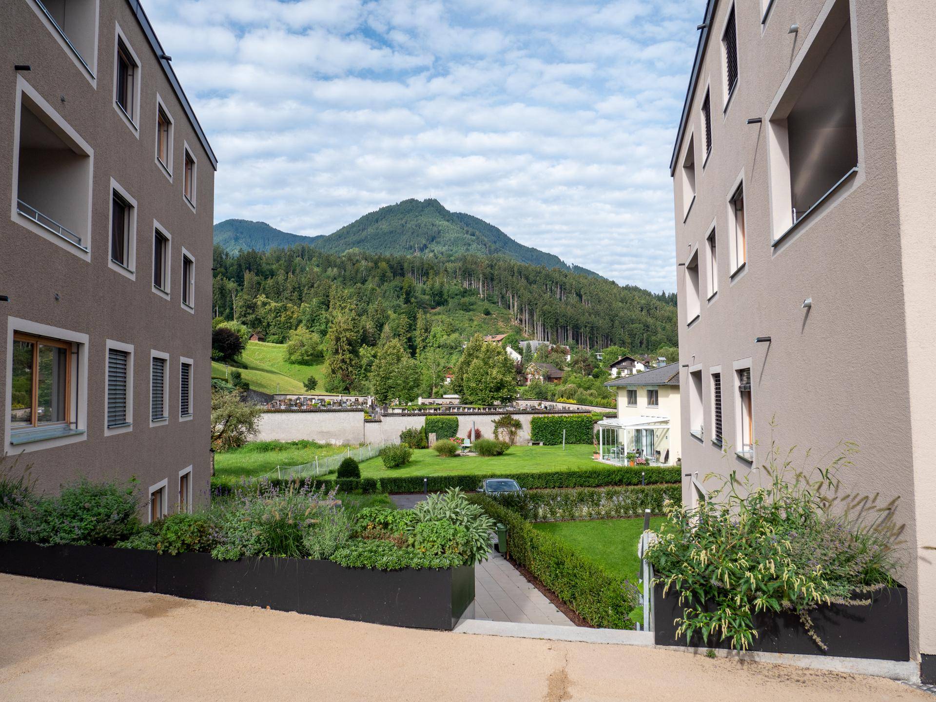 Blick zwischen zwei Gebäuden auf eine gepflegte Gartenanlage mit Hecken und bewaldeten Bergen im Hintergrund.