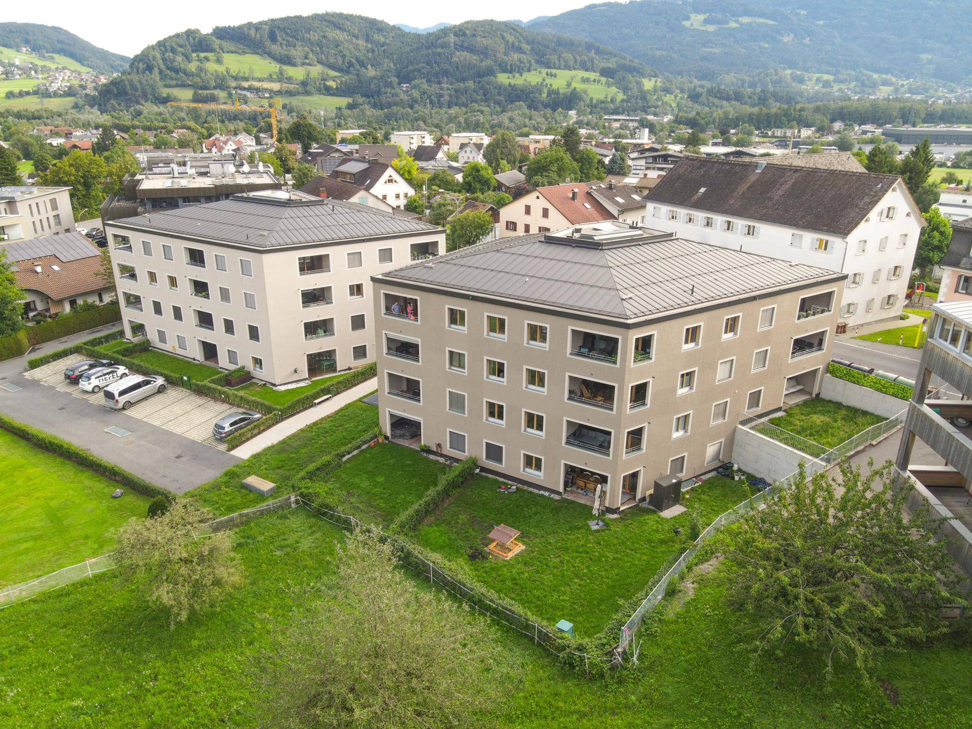 Luftaufnahme zweier moderner Wohngebäude mit viel Grünfläche und einem Spielplatz, umgeben von Bergen.