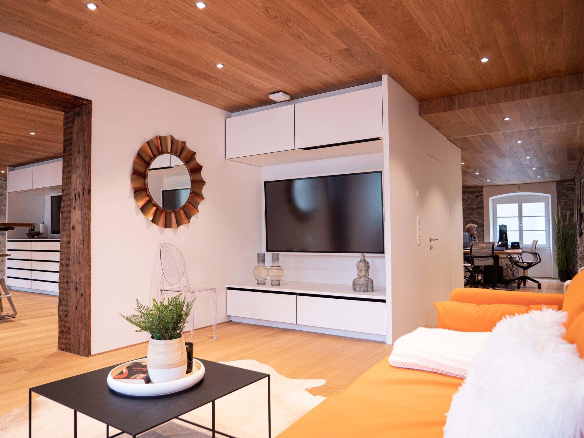 Wohnzimmer mit Wandfernseher, dekorativem Spiegel und Blick auf den angrenzenden Bürobereich.