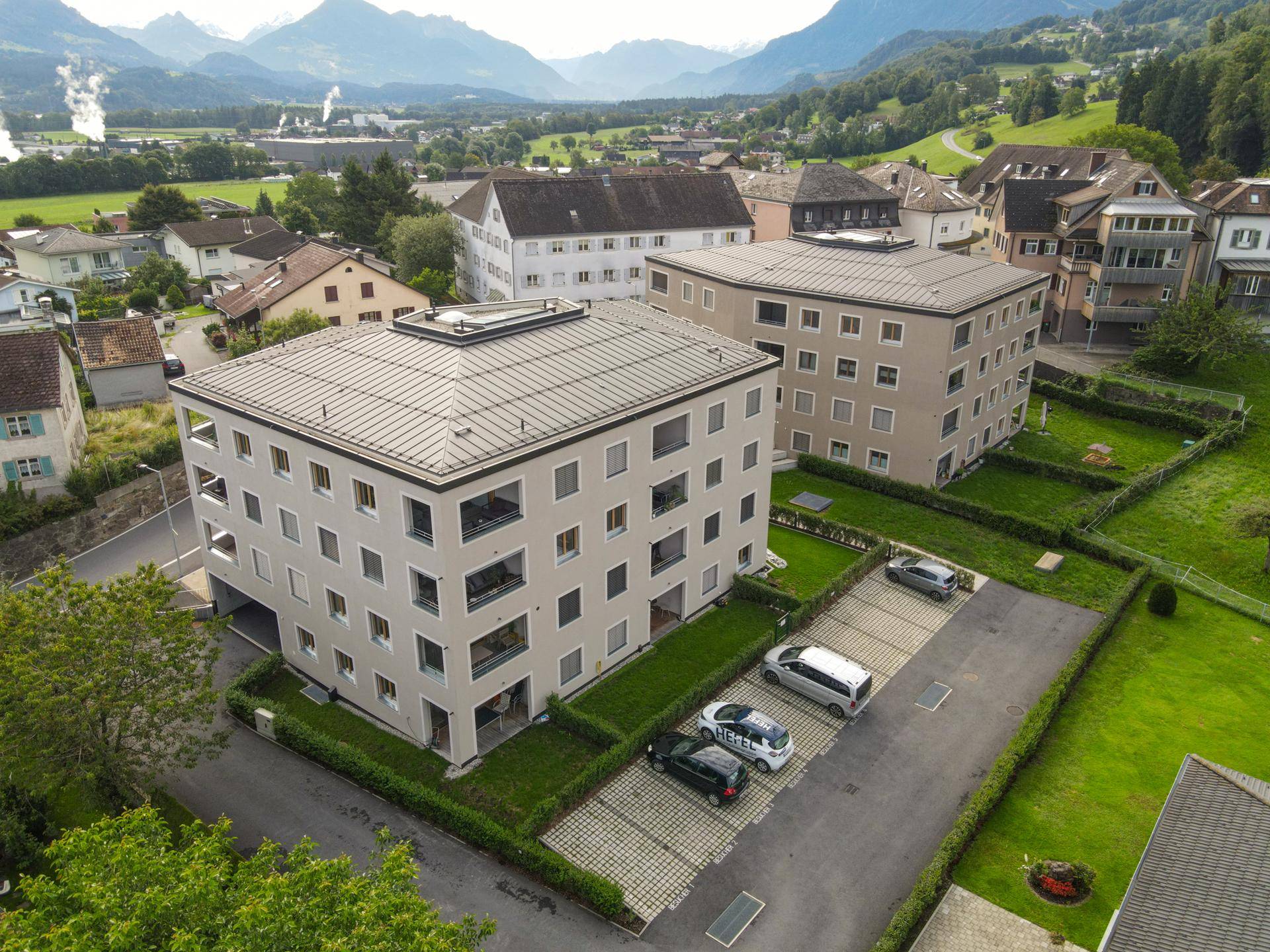 Luftaufnahme von zwei modernen Wohngebäuden mit Flachdächern, umgeben von Grünflächen und Parkplätzen, mit Bergblick.