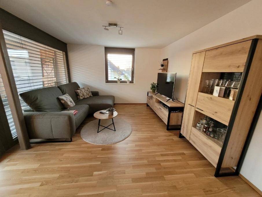 Gemütliches Wohnzimmer mit Sofa, Fernseher und modernem Holzschrank, hell durch große Fenster.