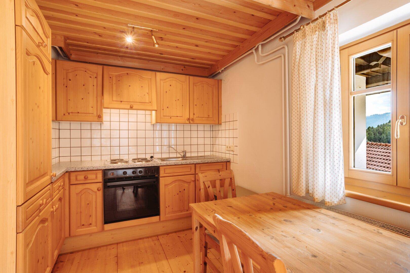 Kleine Küche mit Holzschränken, Esstisch und einem Fenster mit Blick ins Grüne.