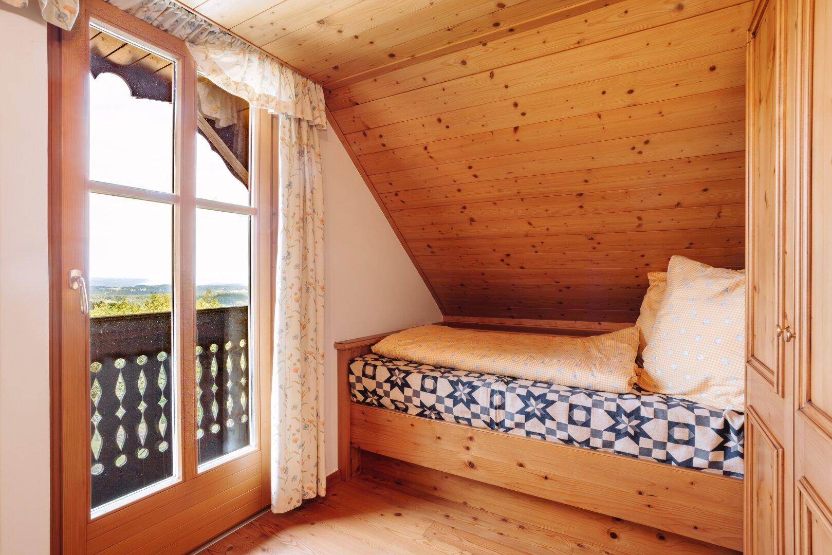 Kleines Schlafzimmer mit Einzelbett, schräger Holzdecke und direktem Zugang zum Balkon.