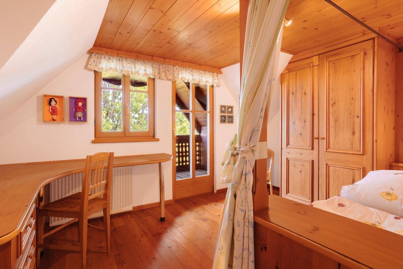 Schlafzimmer mit Schreibtisch, Holzschrank, schräger Holzdecke und Balkonzugang.