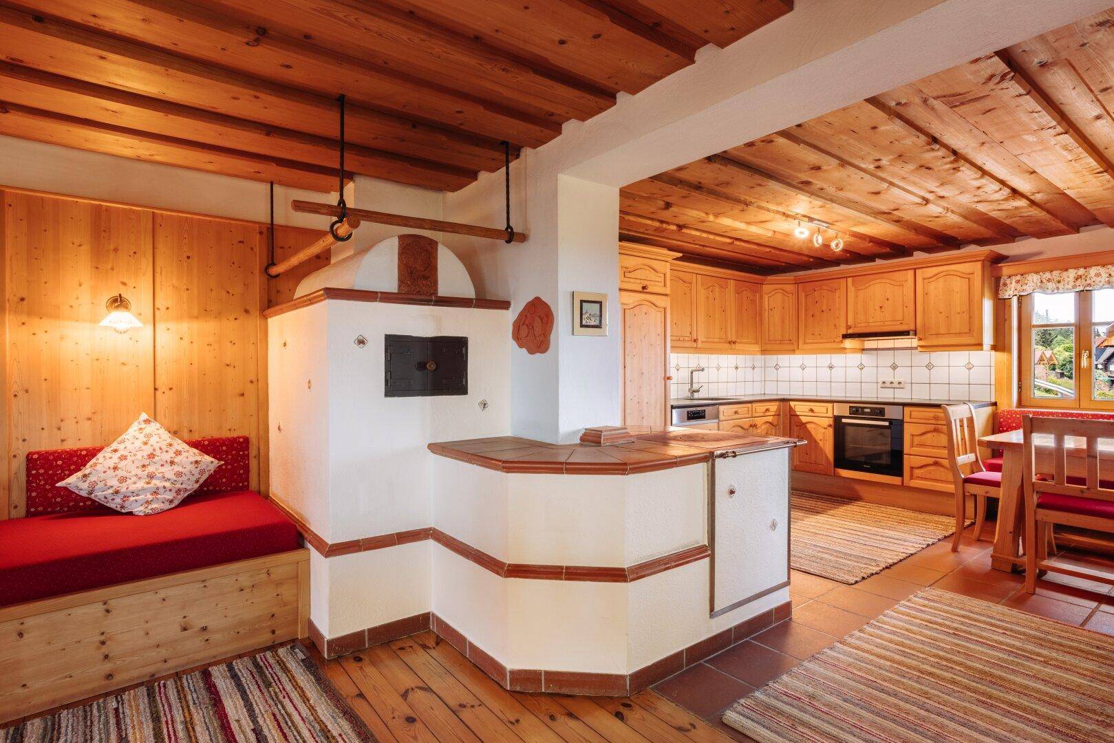 Offener Wohnbereich mit traditionellem Kachelofen, Holzbalkendecke und integrierter Küche im Hintergrund.