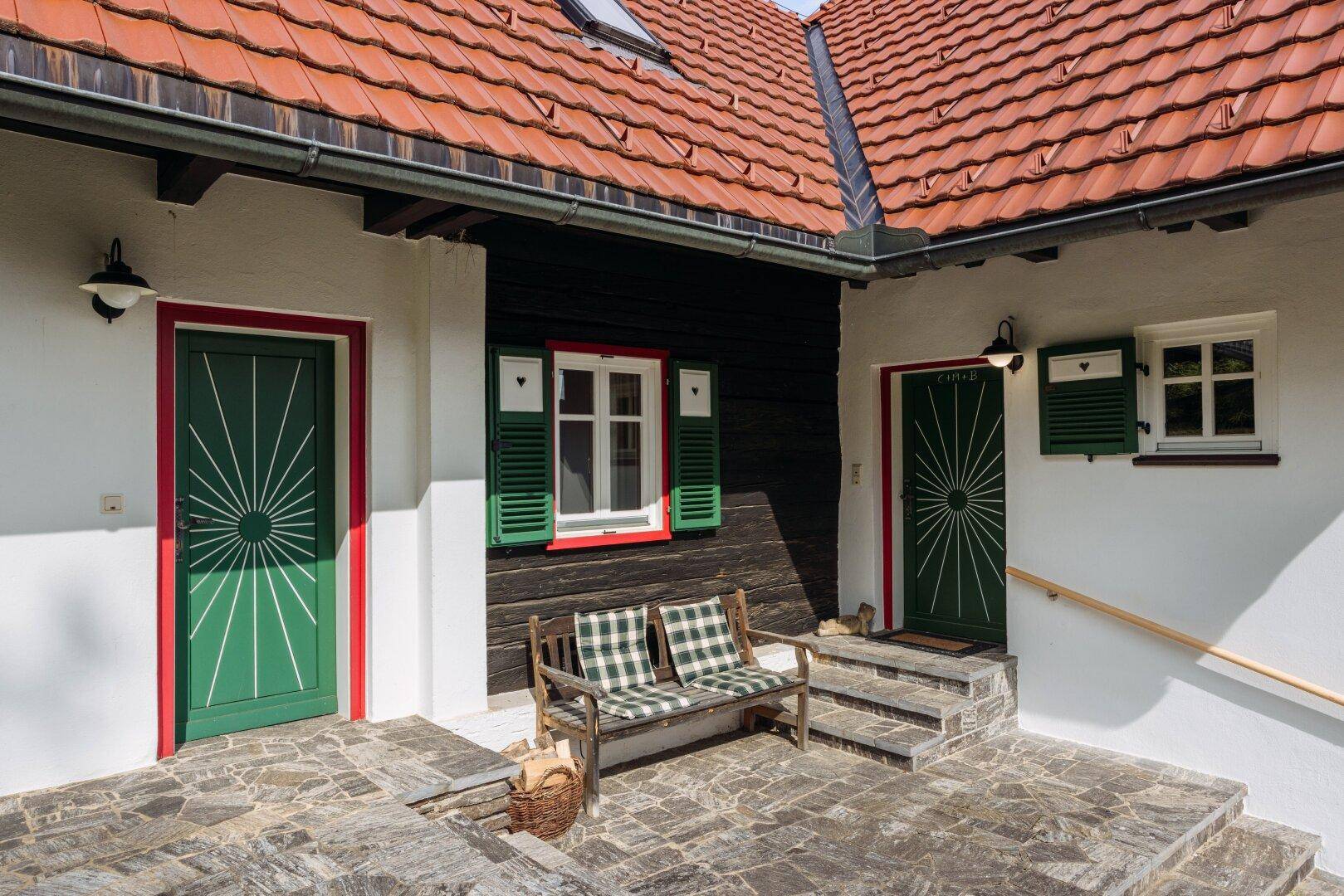 Eingangsbereich des Hauses mit zwei grünen Türen, einer Holzbank und rustikalem Steinboden.
