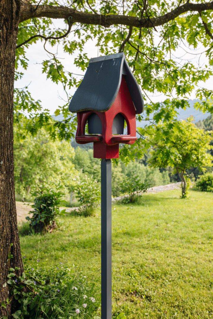 Rotes Vogelhäuschen auf einem Pfahl in einem weitläufigen grünen Garten mit Bäumen.