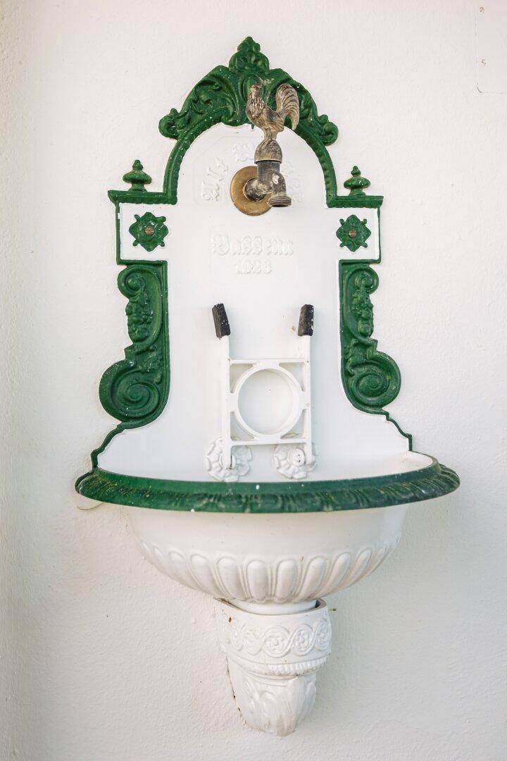 Dekorativer Wandbrunnen mit Hahn und grünen Ornamenten an einer weißen Außenwand.