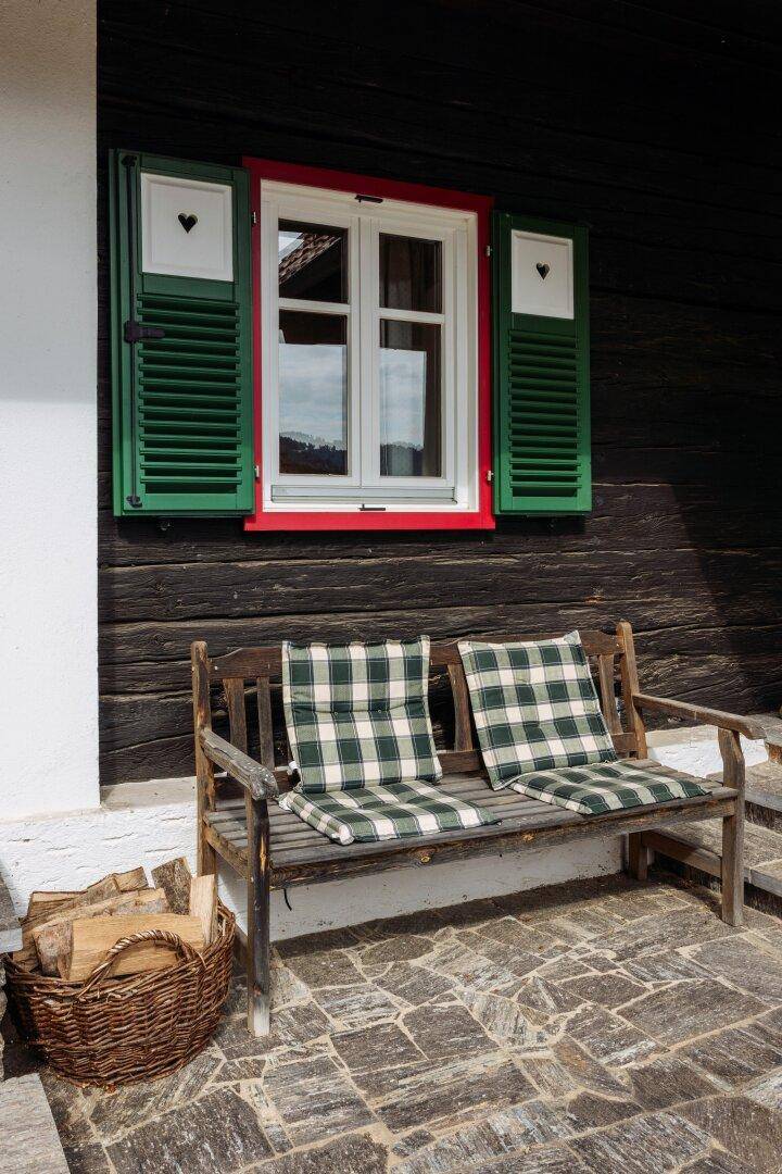 Rustikale Holzbank mit karierten Kissen vor einem Fenster mit grünen Fensterläden.