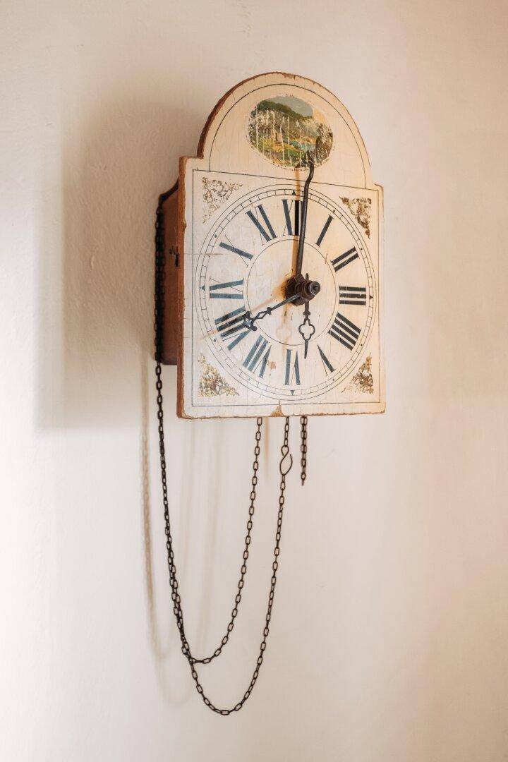 Alte Wanduhr mit römischen Ziffern und Kettenwerk, an einer hellen Wand montiert.
