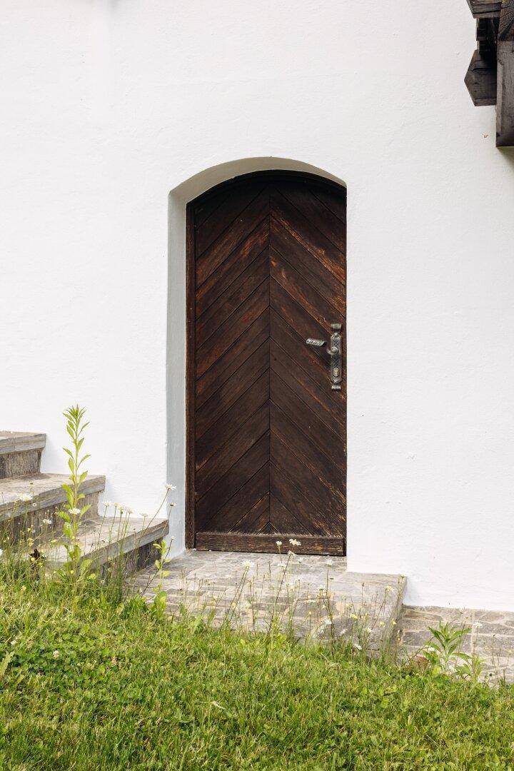 Rustikale Holztür mit Rundbogen und schmiedeeisernem Griff in einer weißen Fassade.