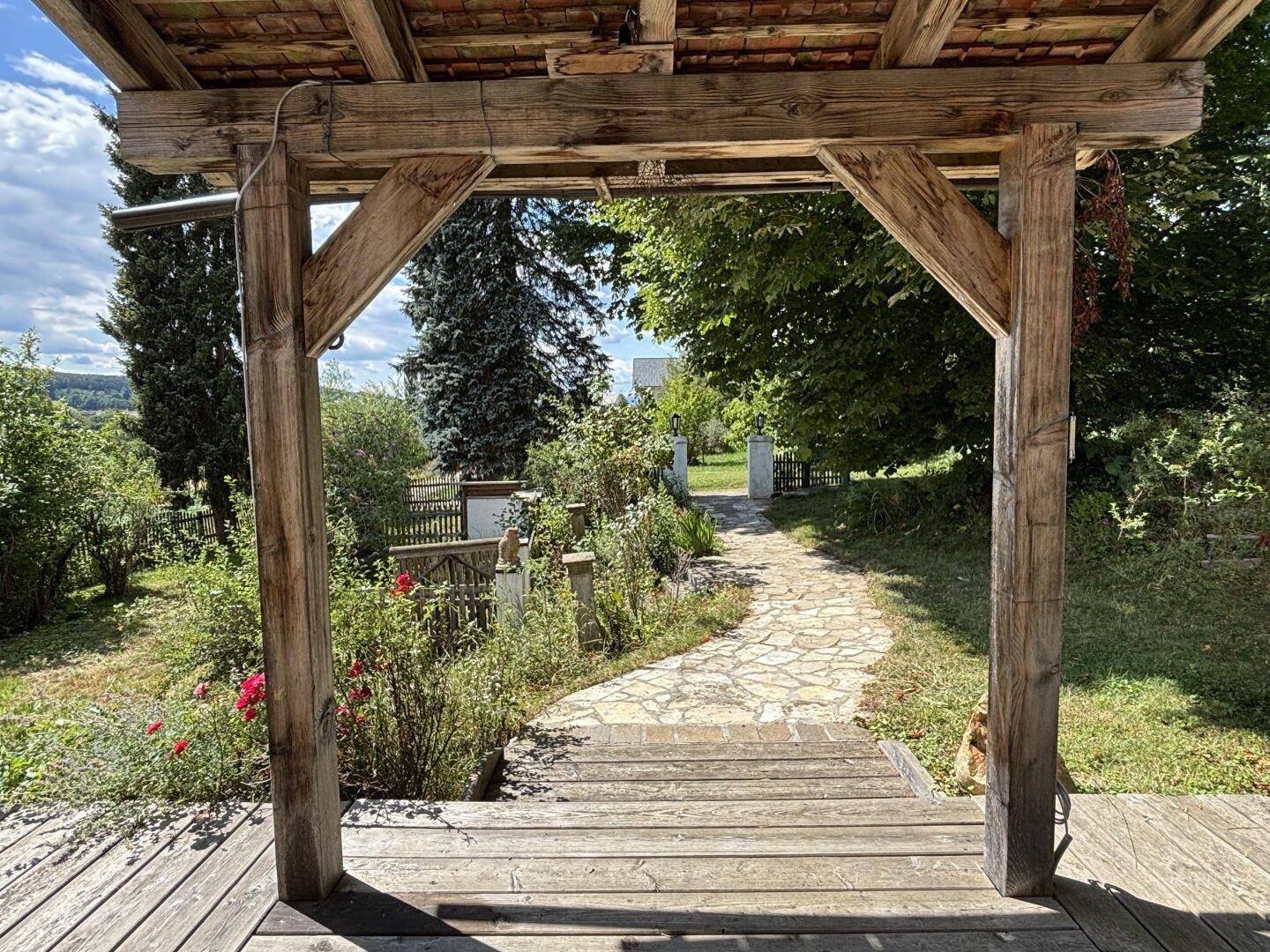 Rustikale Holzpergola am Eingang eines Gartens mit Steinweg und üppiger Bepflanzung.