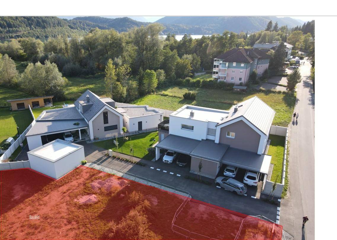 Luftaufnahme eines Wohngebiets mit modernen Häusern, See im Hintergrund und markiertem Baugrundstück.