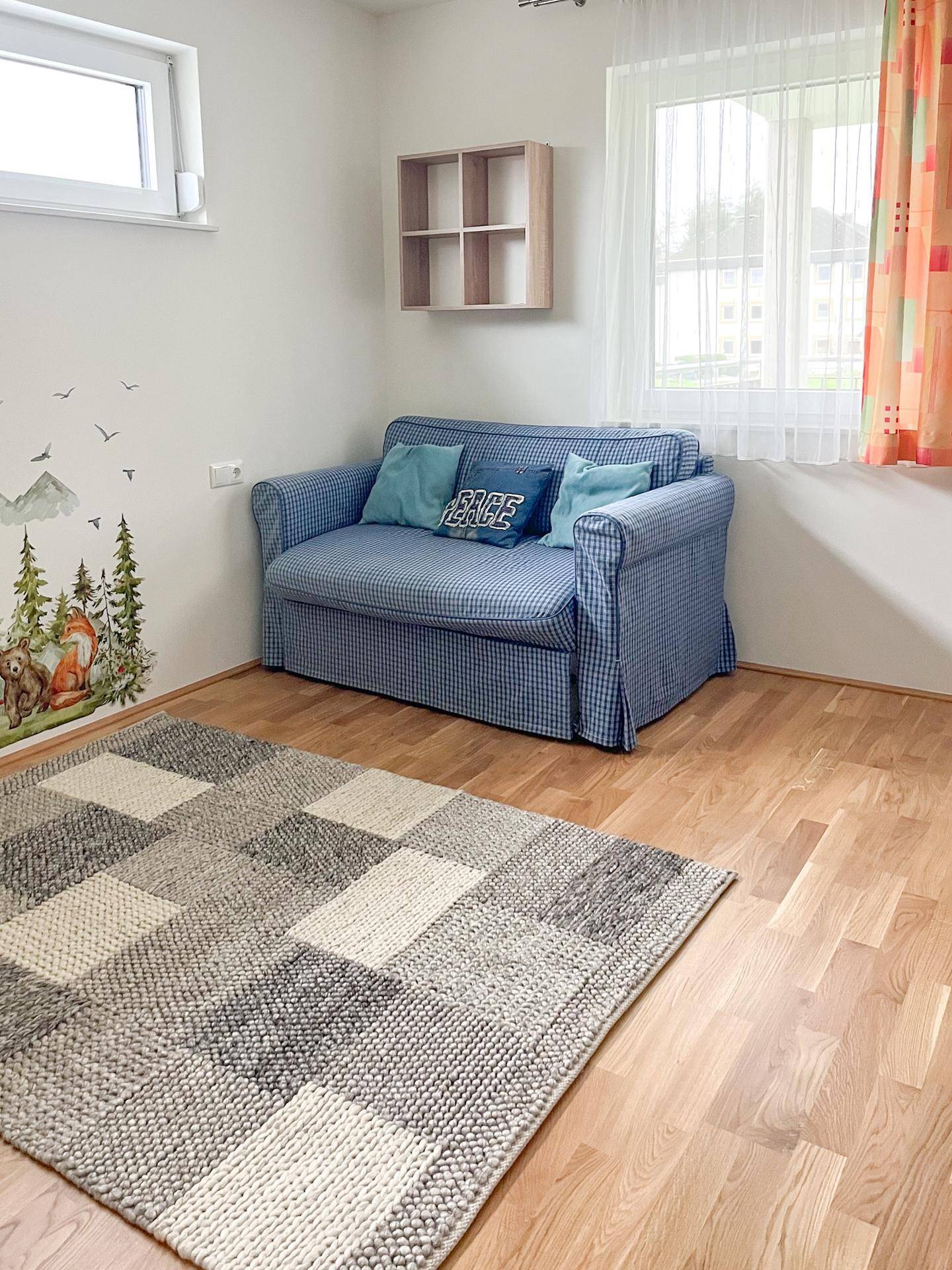 Gemütliches Schlafzimmer mit blauem Schlafsofa, Holzboden und Fenster mit Vorhängen.