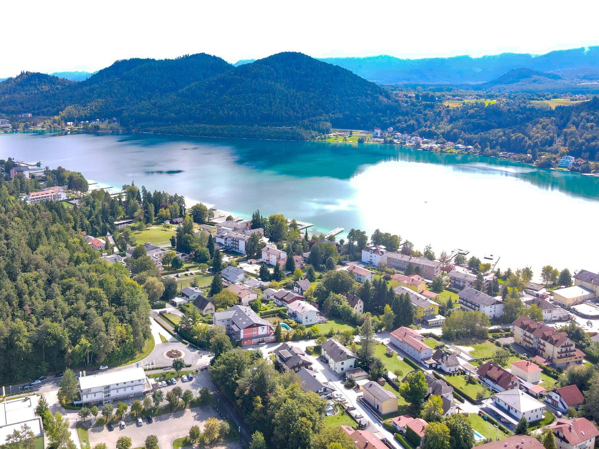 Panoramablick auf einen malerischen See, umgeben von grünen Hügeln, Wäldern und einer Siedlung.