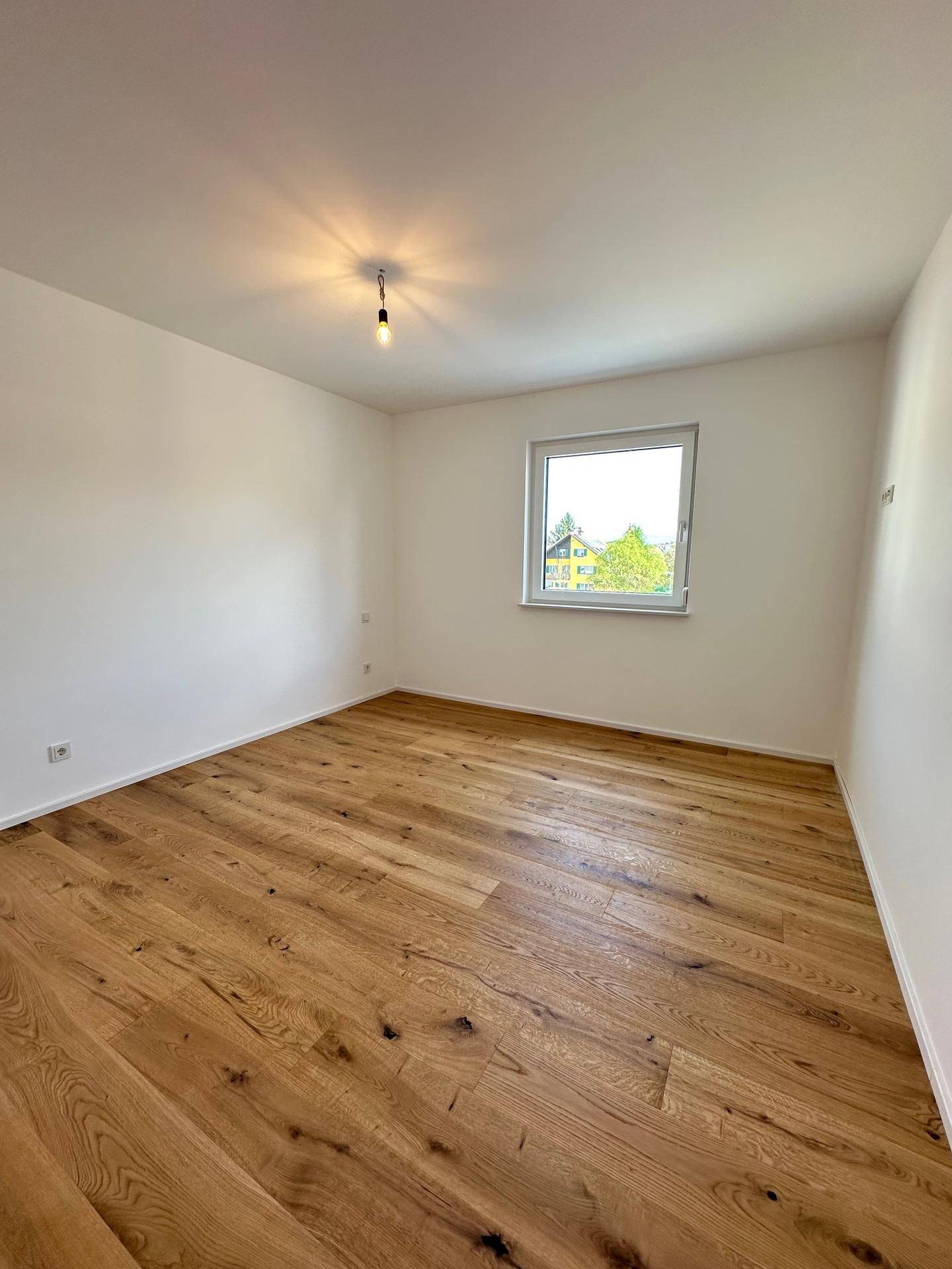 Leeres Schlafzimmer mit Holzboden und Fenster, das viel Tageslicht hereinlässt.