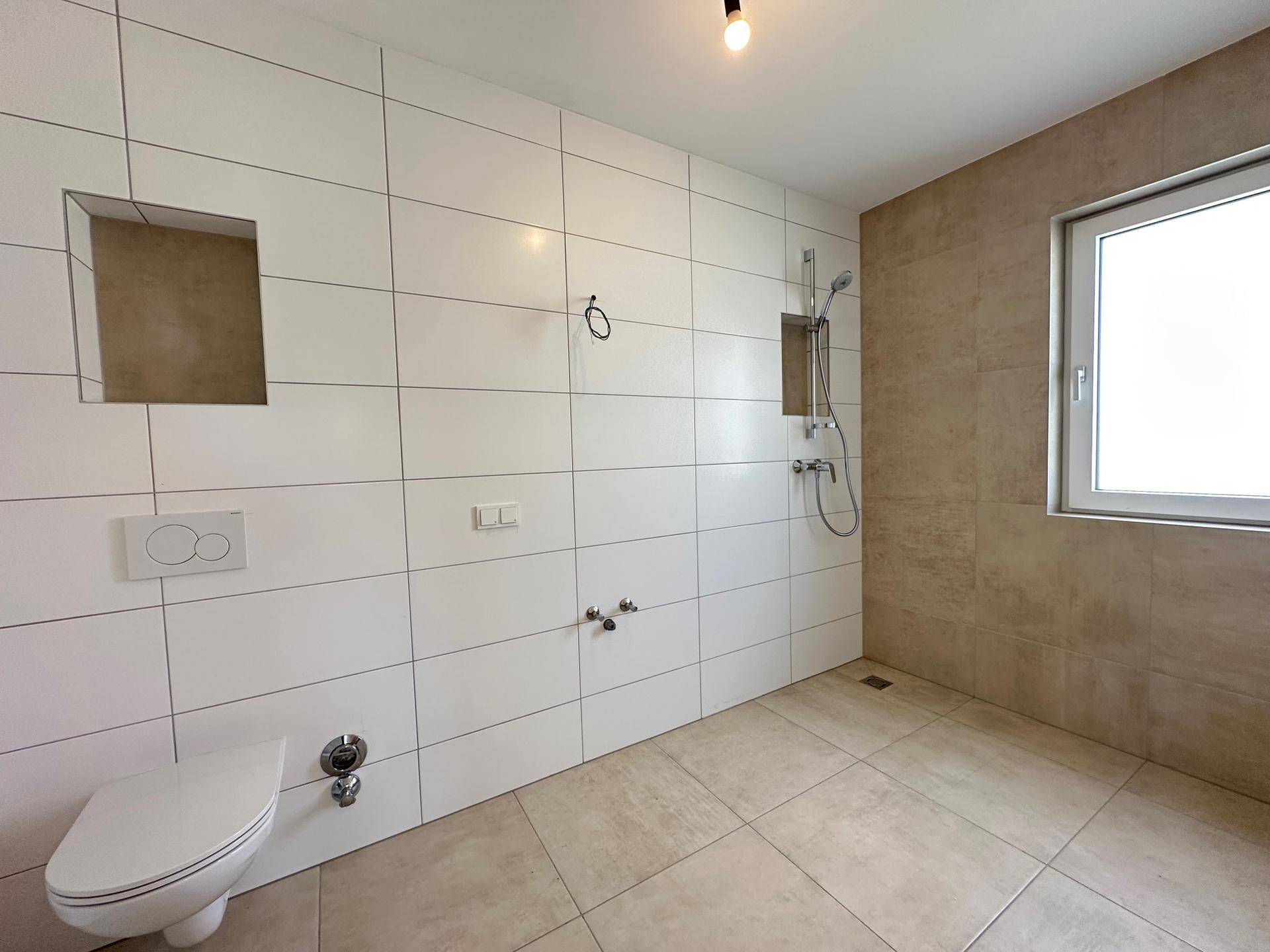 Modernes Badezimmer im Rohbau mit Fliesen, Duschbereich und Wand-WC-Vorbereitung.