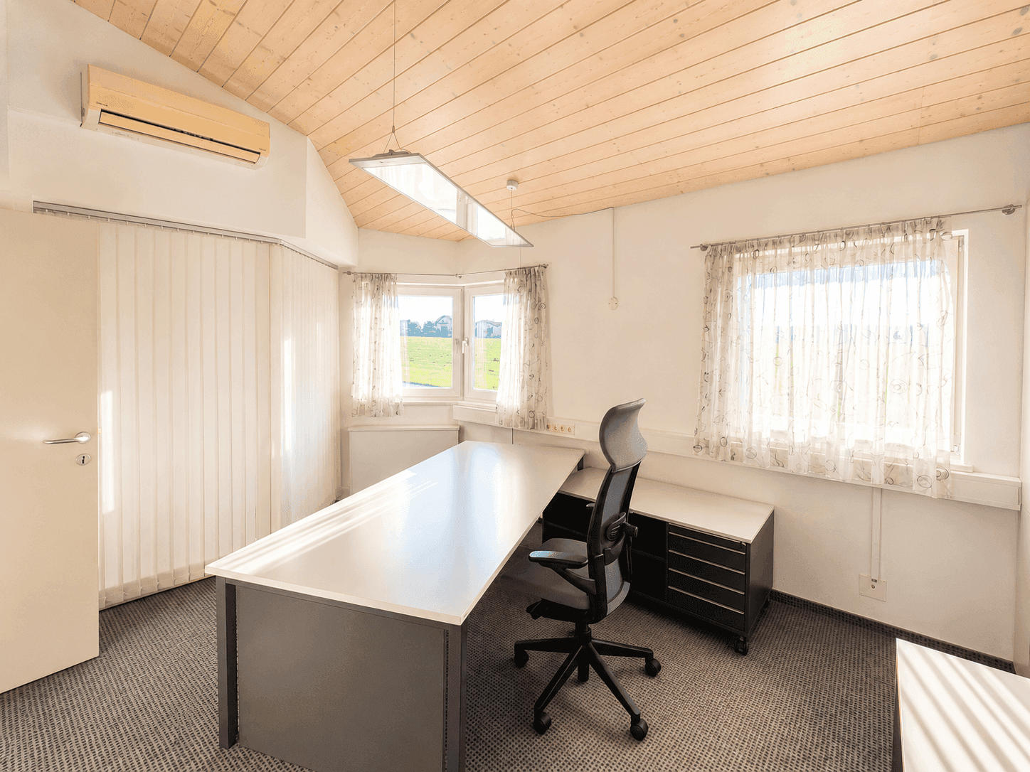 Geräumiges Büro mit Schreibtisch, Bürostuhl und Fenstern mit Blick ins Grüne.